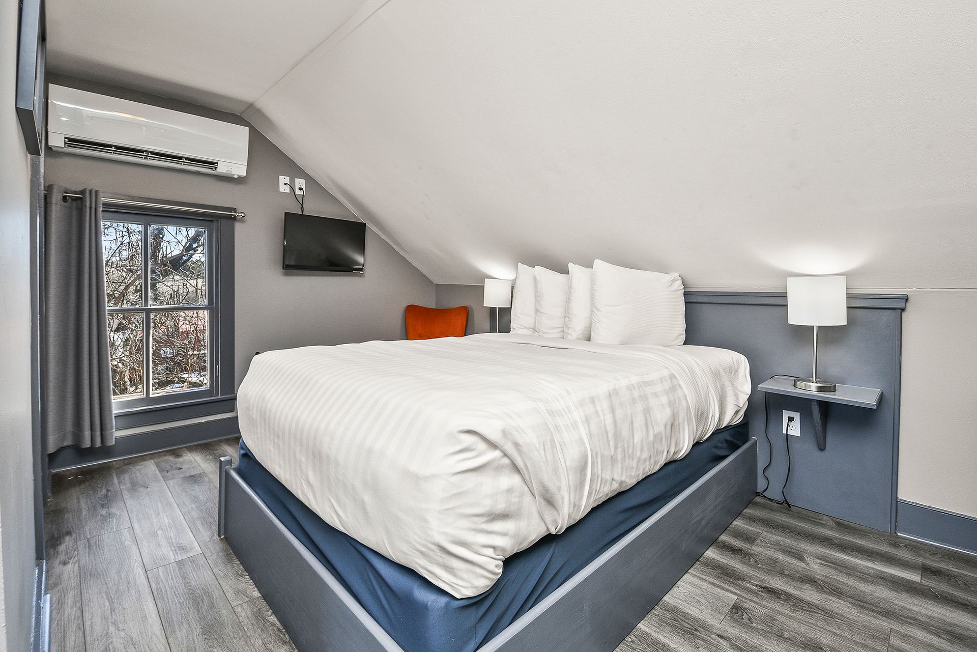 A cozy attic bedroom featuring a white bed with a blue base, a small wall-mounted table with a lamp, and a window.