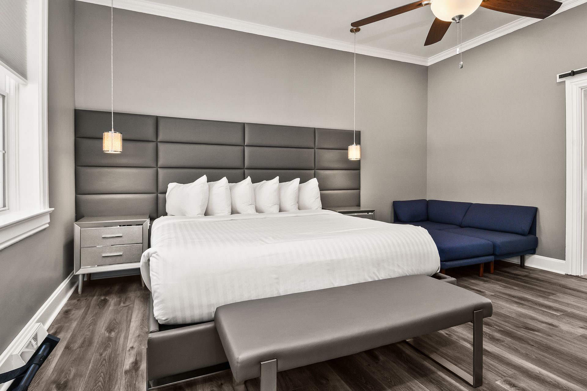 A modern hotel room featuring a king bed with a tall grey headboard, a blue sofa, and a grey bench on wood-look flooring.