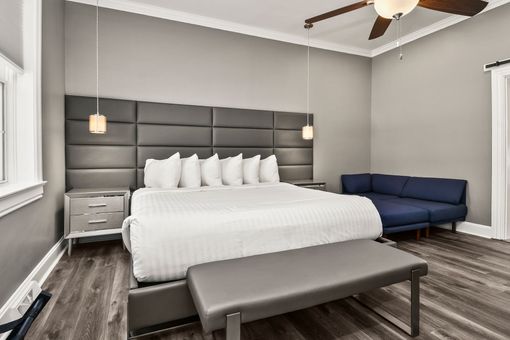 A modern hotel room featuring a king bed with a tall grey headboard, a blue sofa, and a grey bench on wood-look flooring.