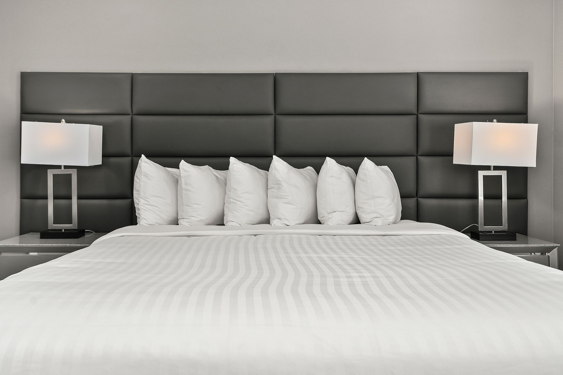 A made bed with crisp white bedding and a row of pillows against a dark, tufted leather headboard with two bedside lamps.