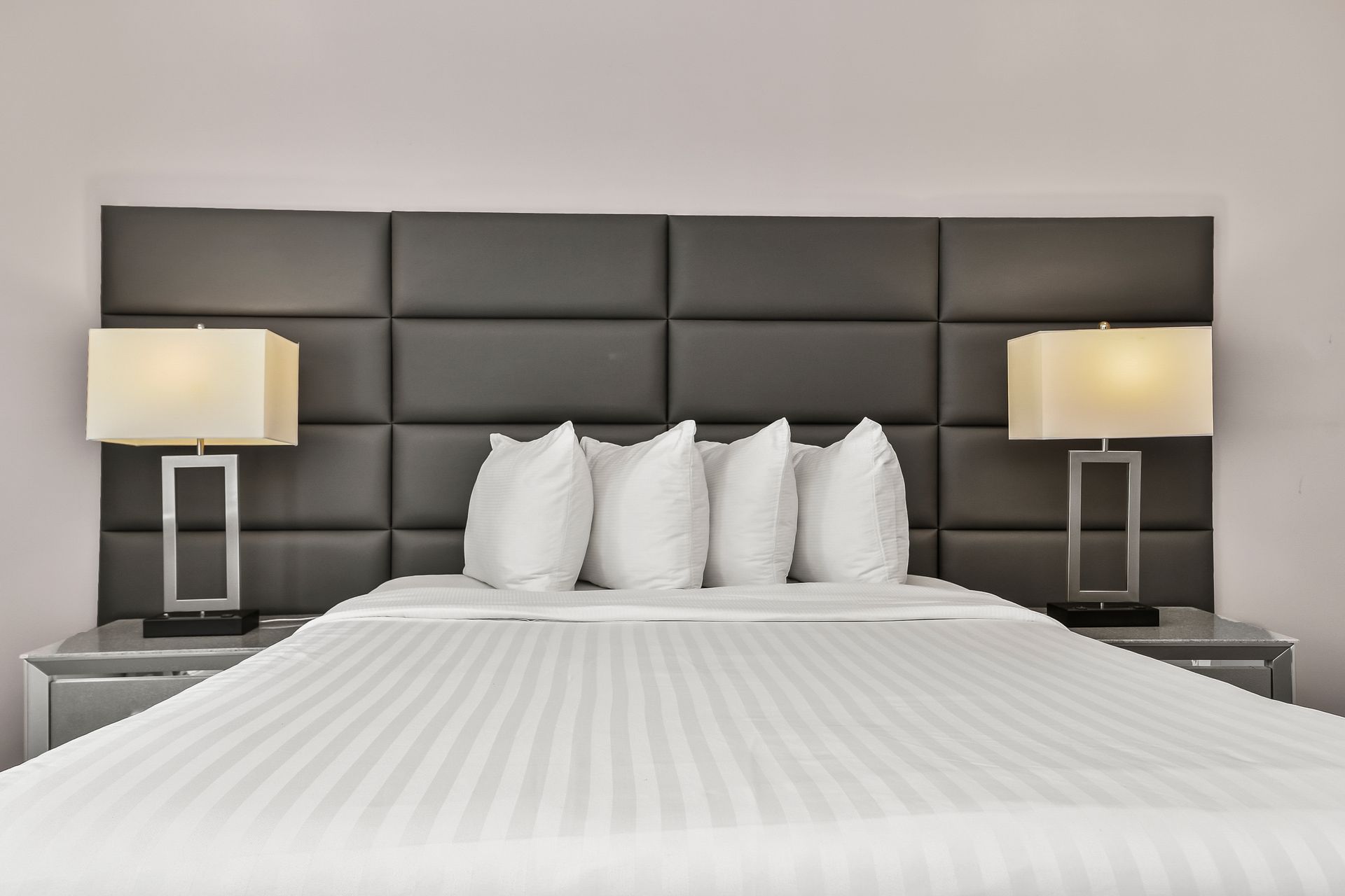 A modern hotel bed with white striped bedding, four pillows, and a large, dark-grey tufted headboard with two table lamps.