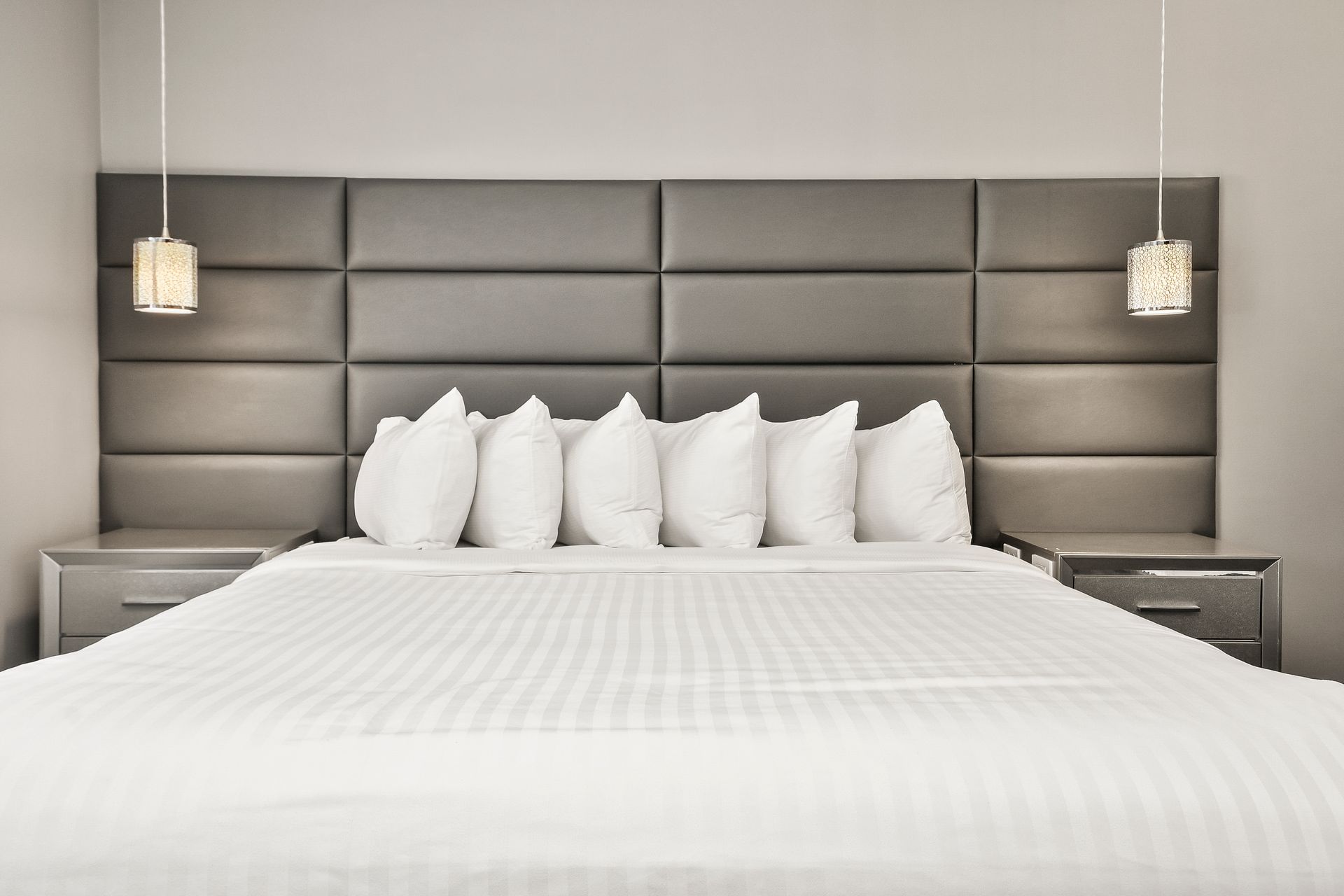 A neatly made bed with white linens and six pillows against a tufted gray headboard with two bedside lamps.