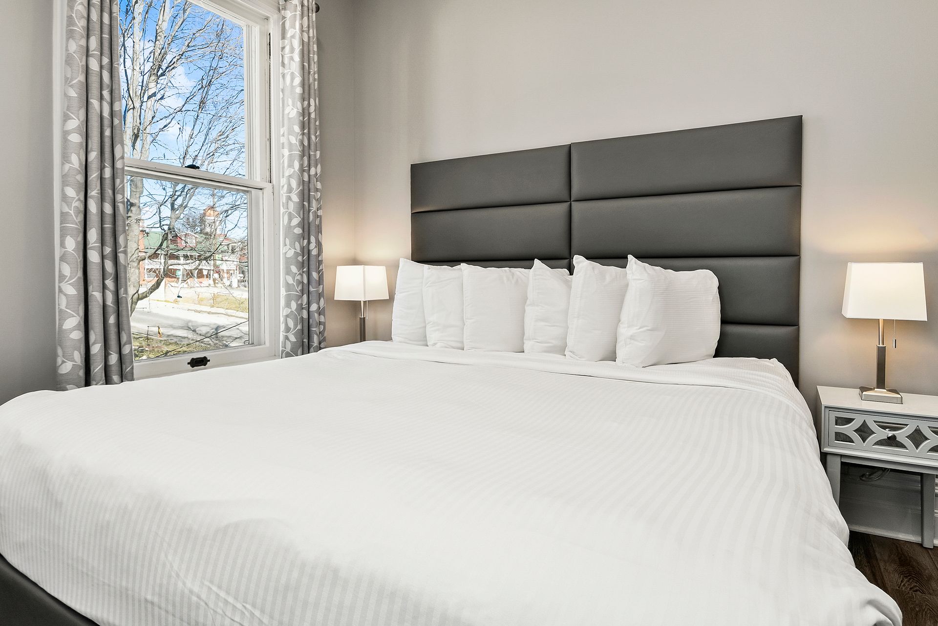 A bedroom featuring a large bed with white bedding, a dark leather headboard, and a window with gray patterned curtains.