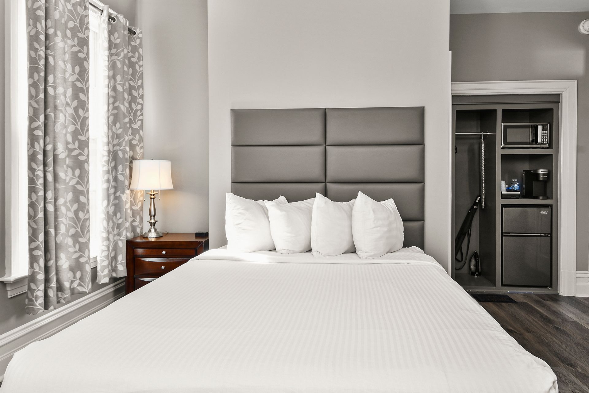 A hotel room with a neatly made bed, grey upholstered headboard, a side table with a lamp, and a recessed kitchenette.