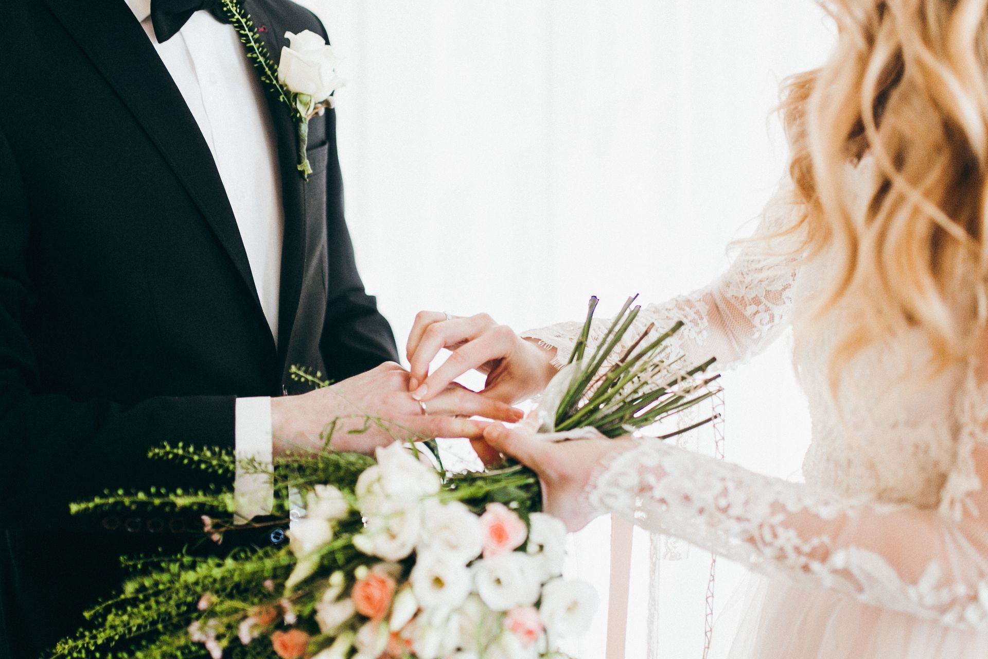 A bride placing a wedding ring on a groom’s finger while holding a floral bouquet.