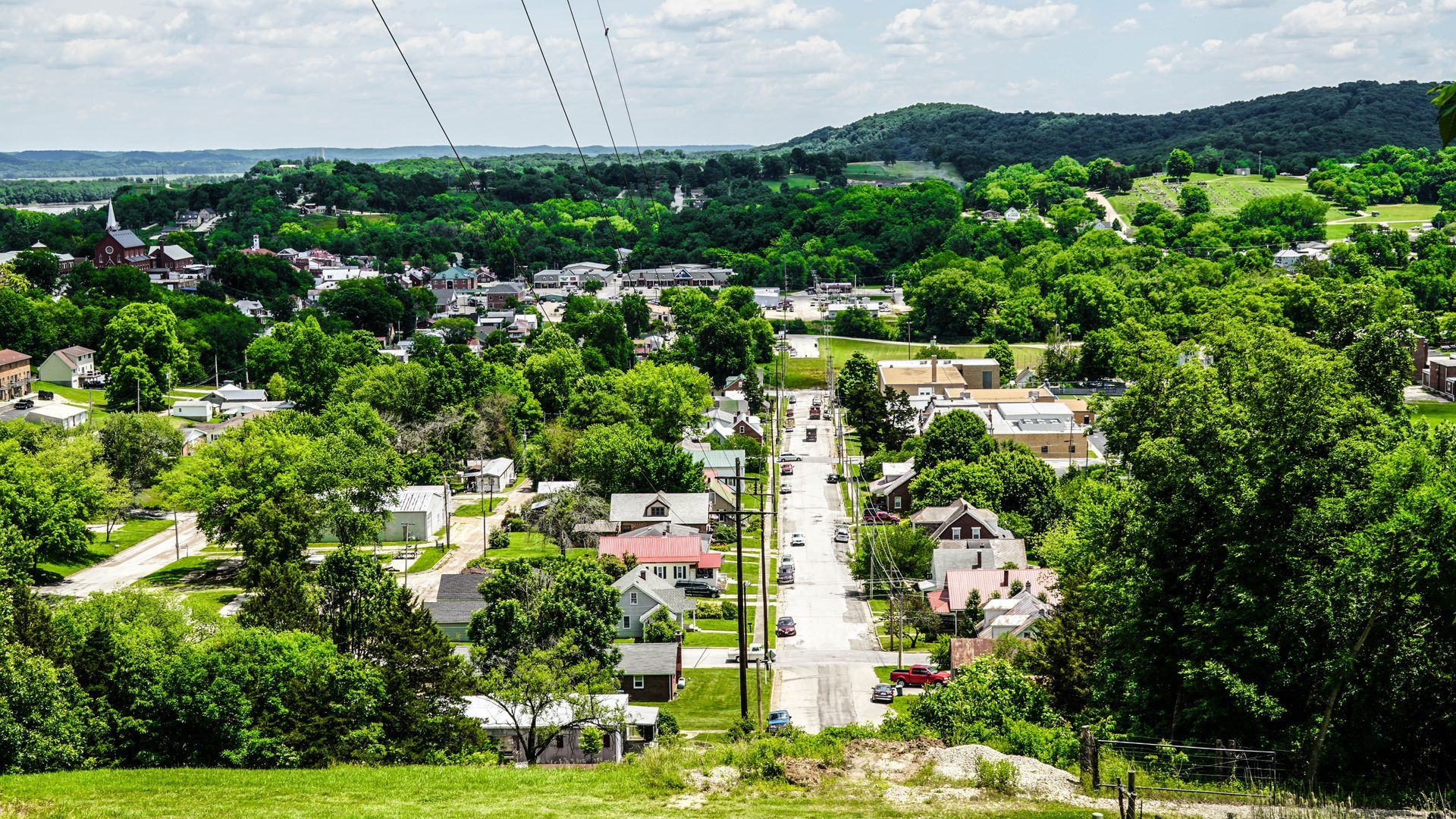 A view of a small town nestled among lush green trees and rolling hills, with a single road leading through the center.