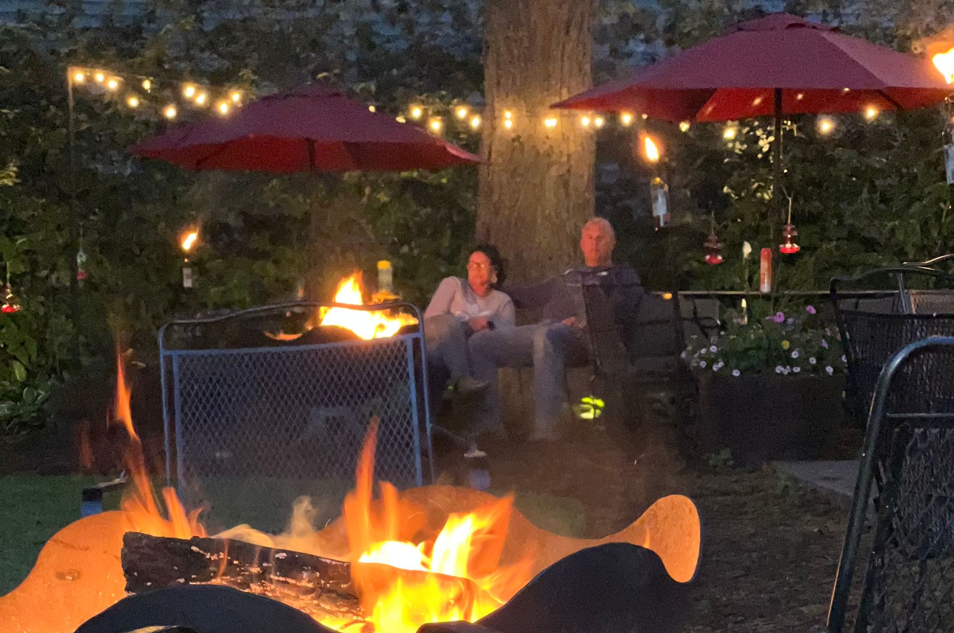 Two people sit on a bench in front of a tree, behind a large fire pit, illuminated by string lights and red umbrellas.