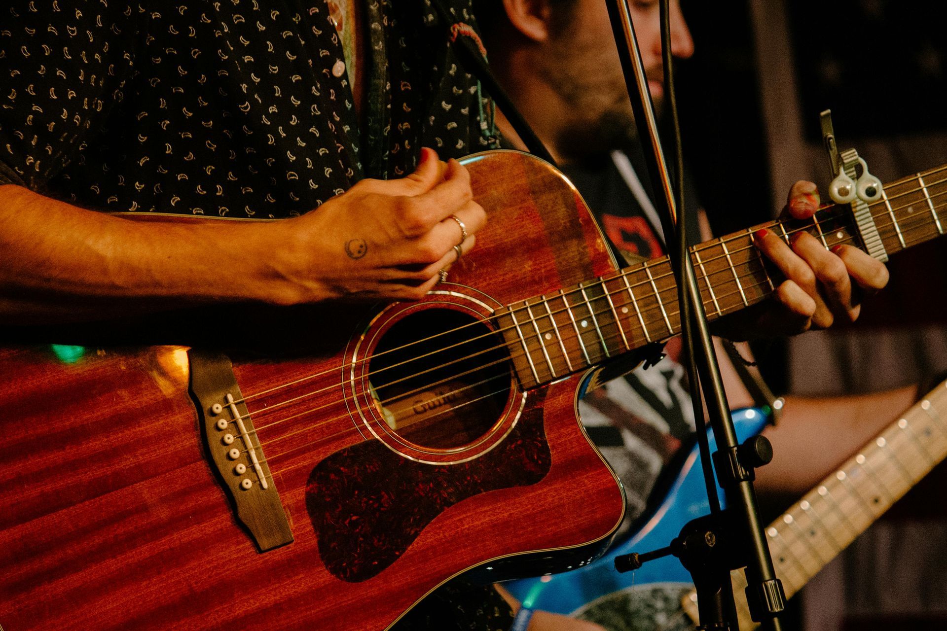 A person plays a reddish-brown acoustic guitar with a capo on the neck, with another musician visible in the background.