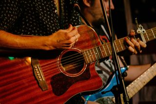 A person plays a reddish-brown acoustic guitar with a capo on the neck, with another musician visible in the background.