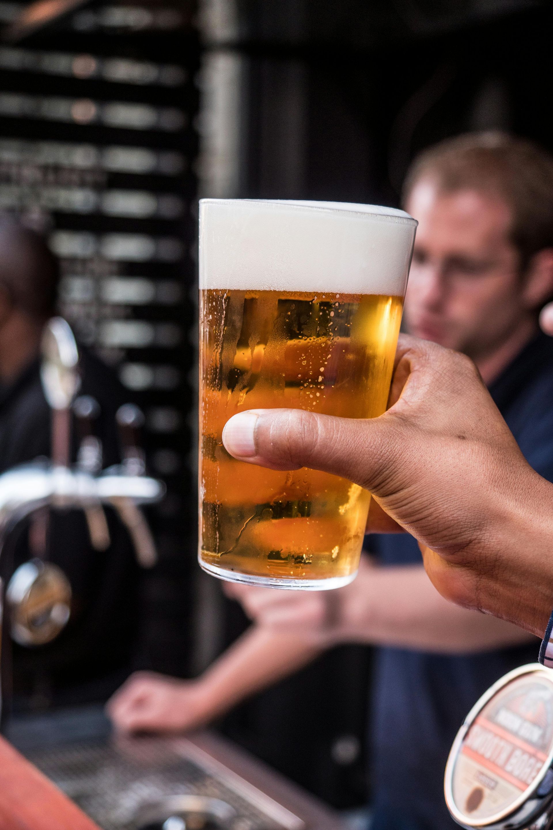 A close-up view of a hand holding a glass of golden beer with foam in a dimly lit bar setting.