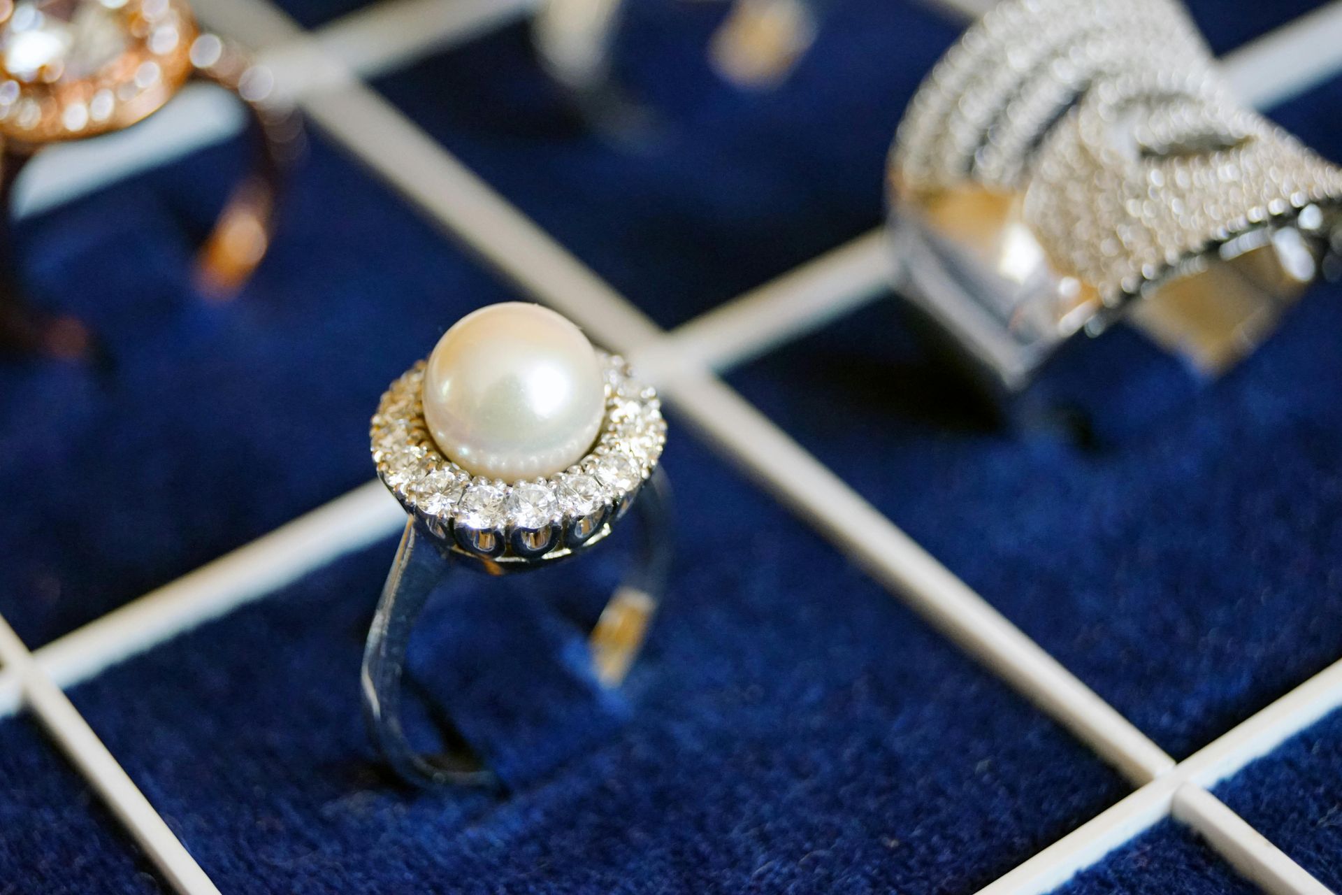 A pearl ring surrounded by small diamonds sits in a blue velvet jewelry organizer next to other rings.