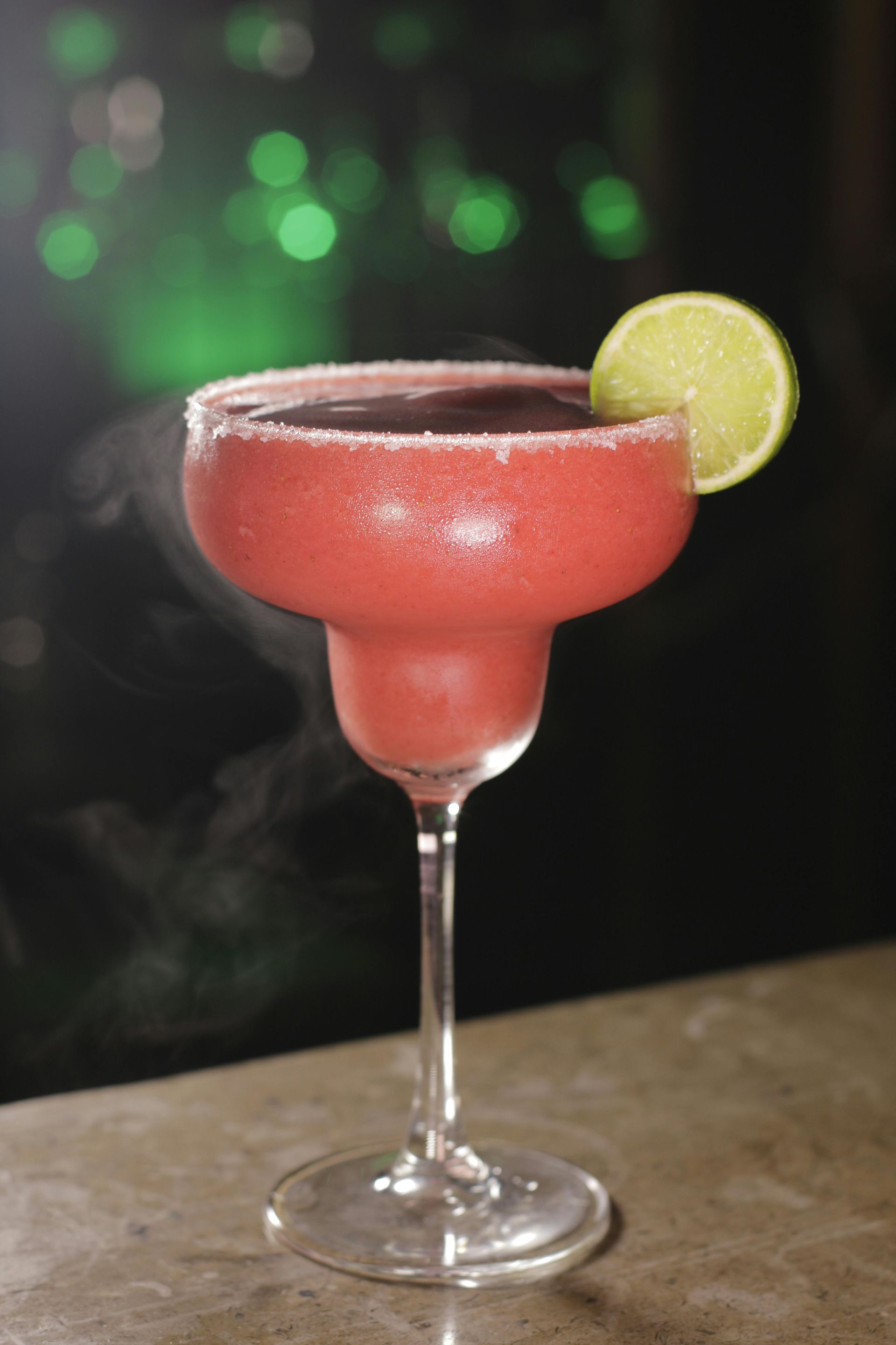 A pink frozen cocktail in a glass with a salted rim and lime garnish, set against a dark background with green bokeh lights.