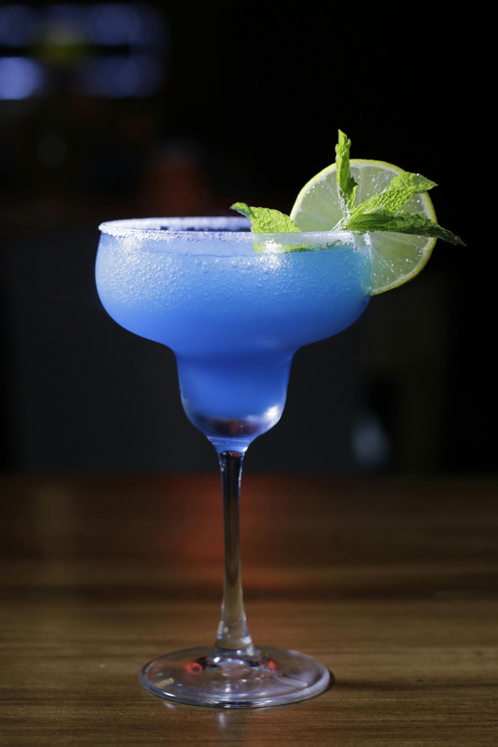 A blue margarita in a stemmed glass with a salted rim, garnished with a lime slice and mint on a wooden table.