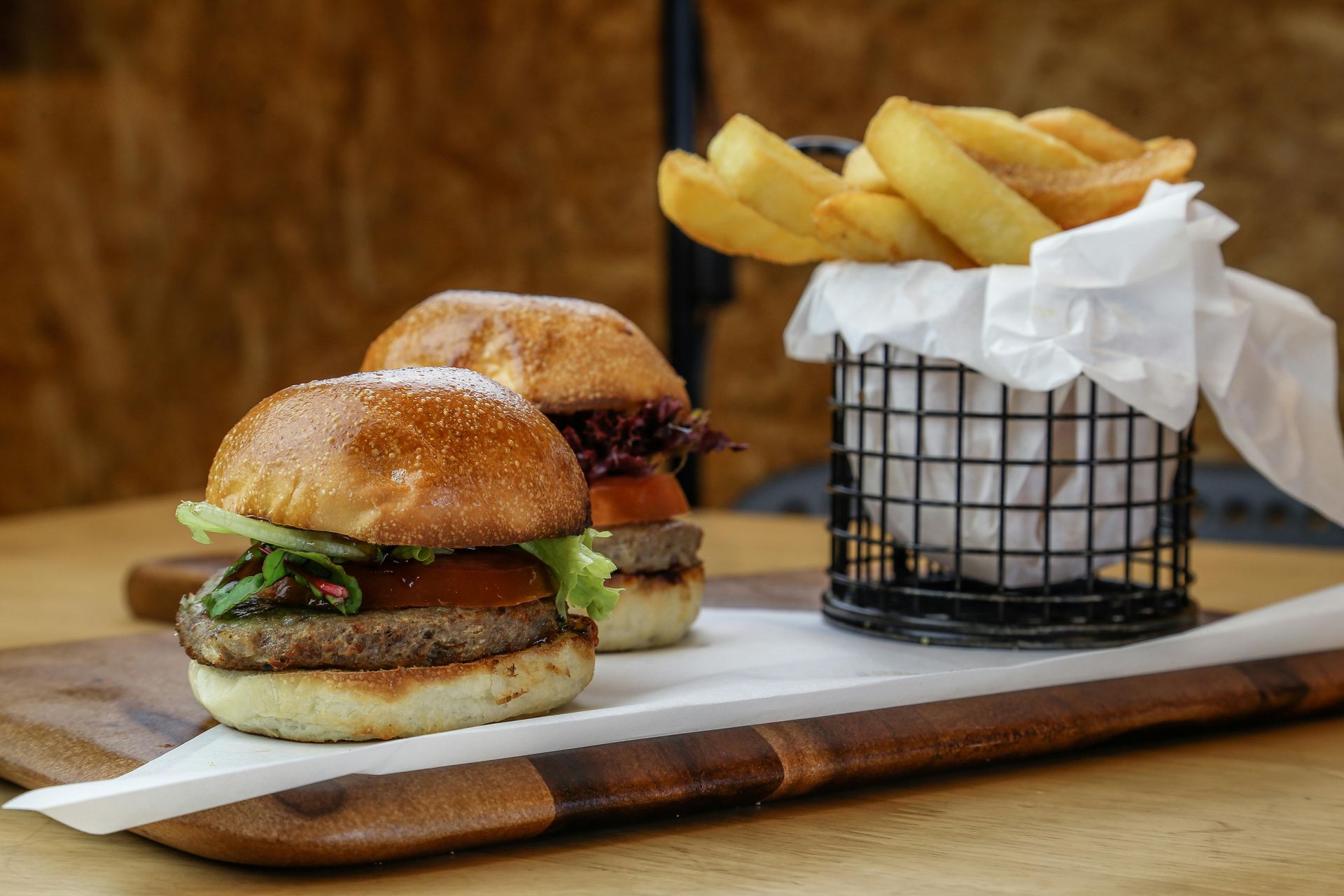 Two gourmet sliders served on a wooden board next to a small metal wire basket filled with thick-cut potato wedges.