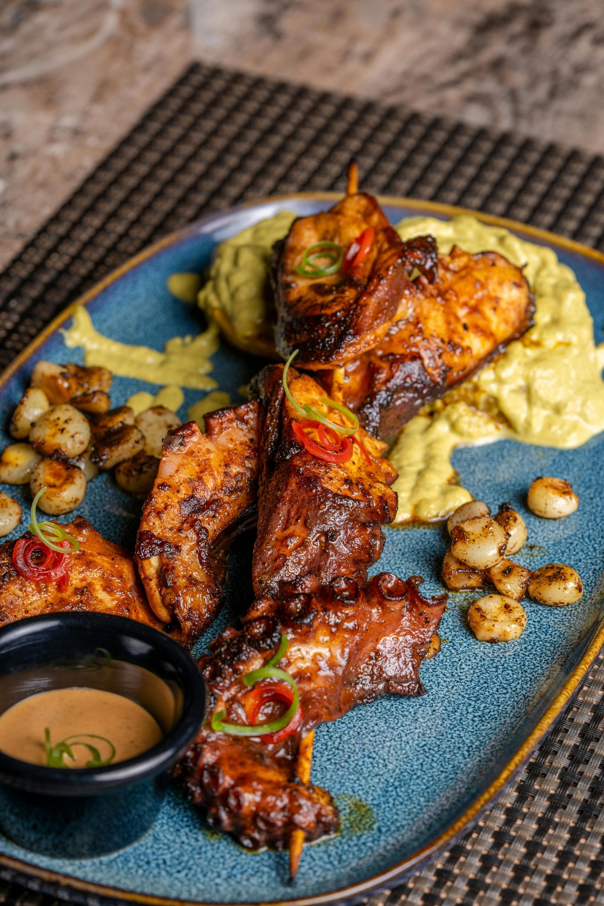 Grilled octopus skewers served on a blue platter with creamy yellow mash, small roasted vegetables, and a dipping sauce.