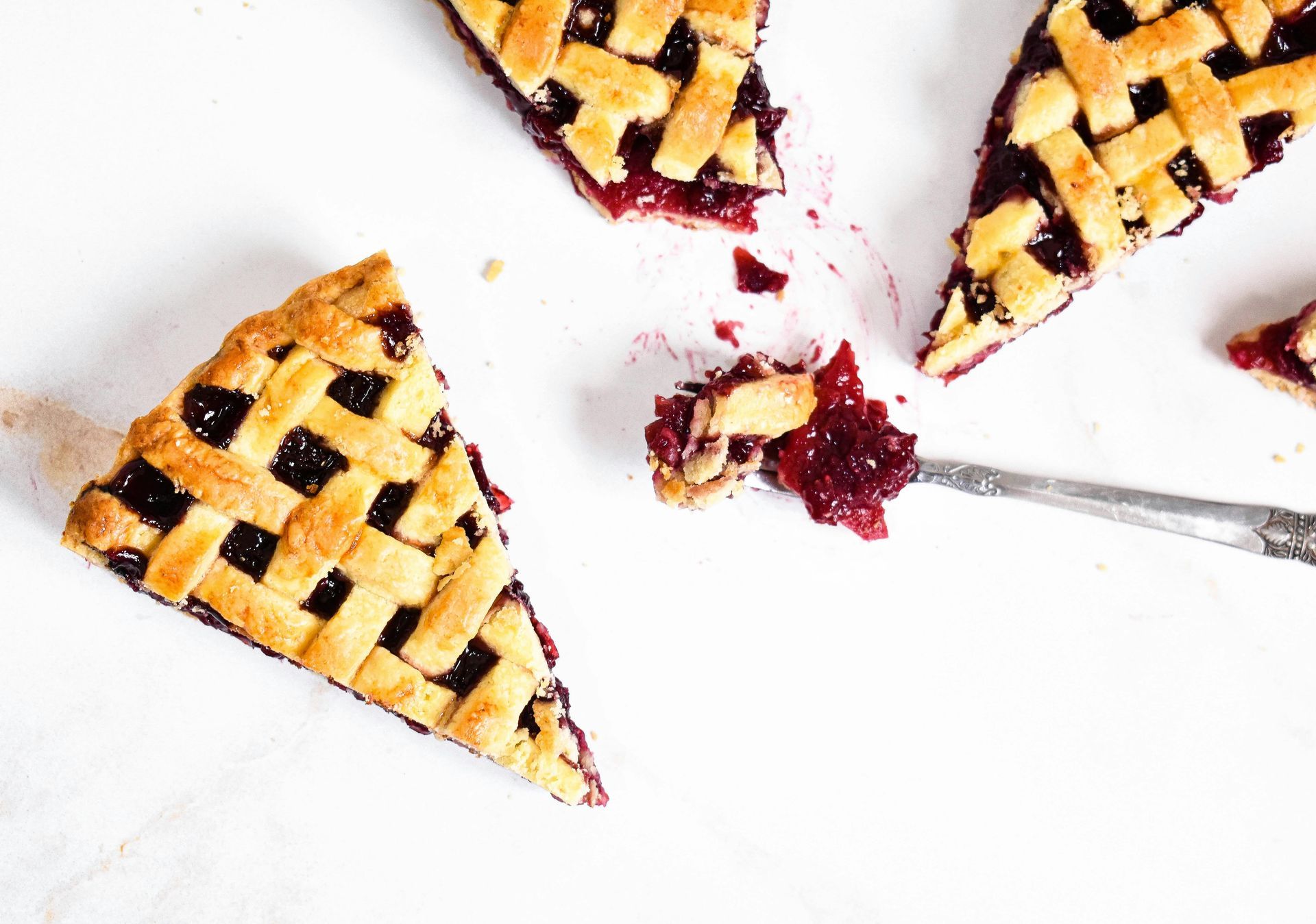 Three triangular slices of lattice-crust berry pie on a white surface, with a fork holding a bite of pie filling.