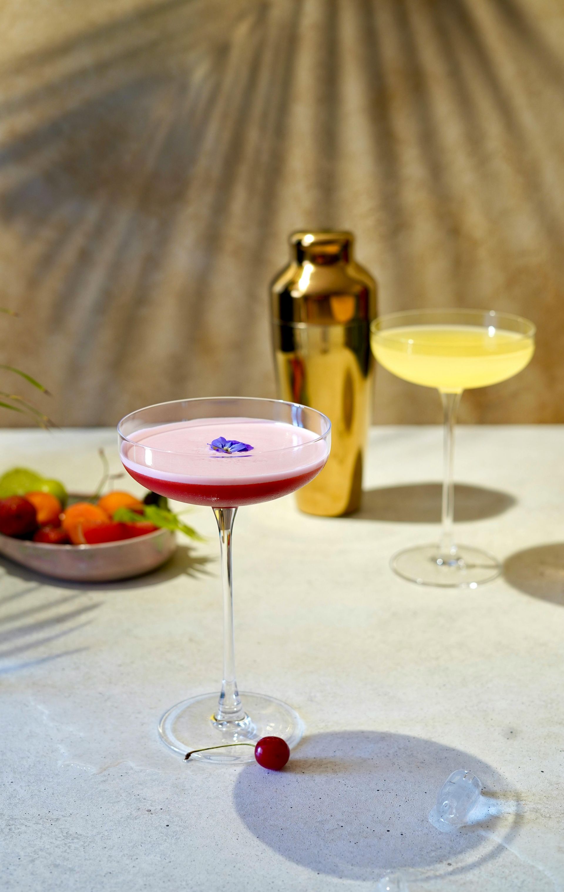 Two colorful cocktails in coupe glasses on a light surface with a gold shaker, bowl of fruit, and palm leaf shadows.