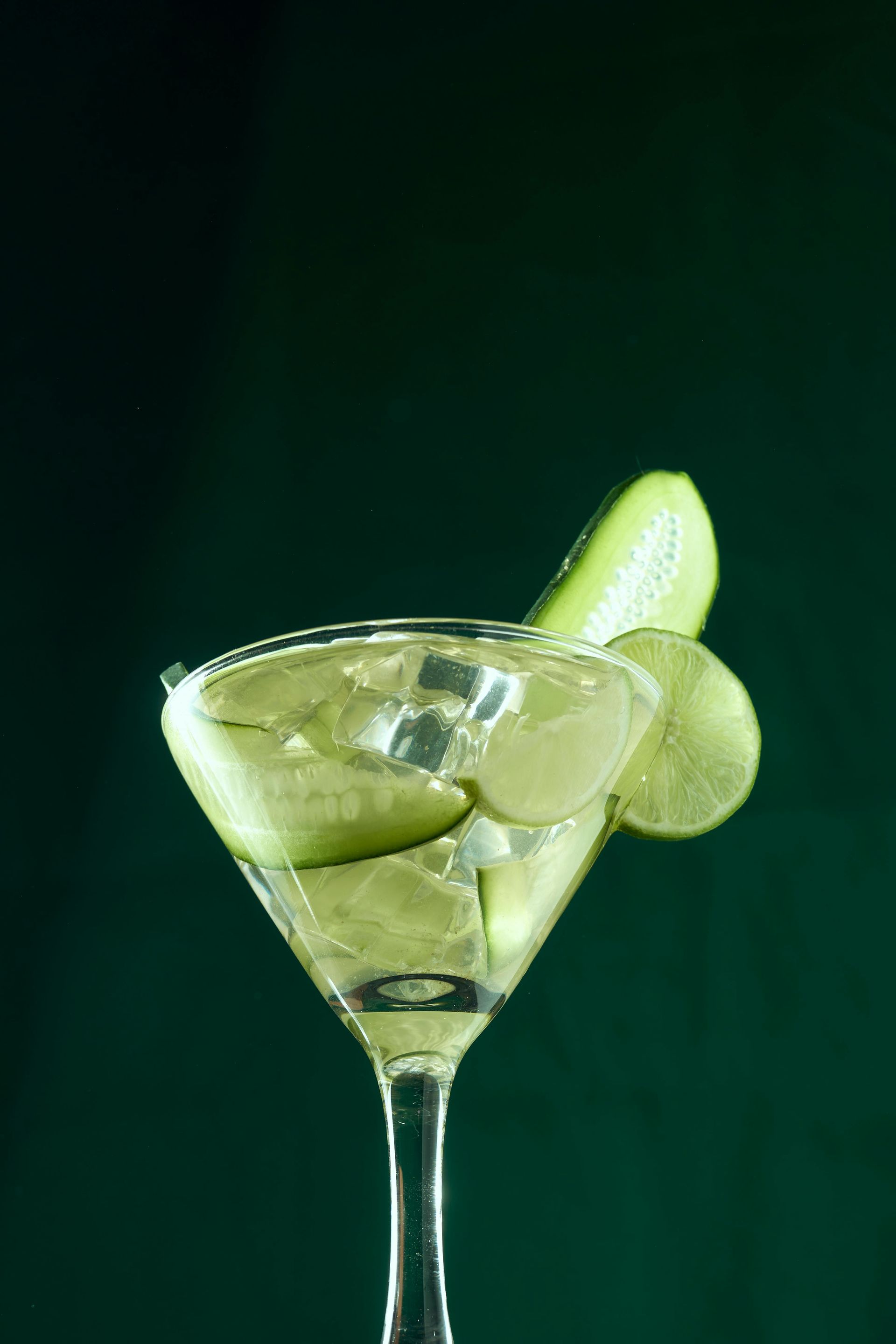 A martini glass filled with ice, cucumber slices, and a lime wheel against a dark green background.