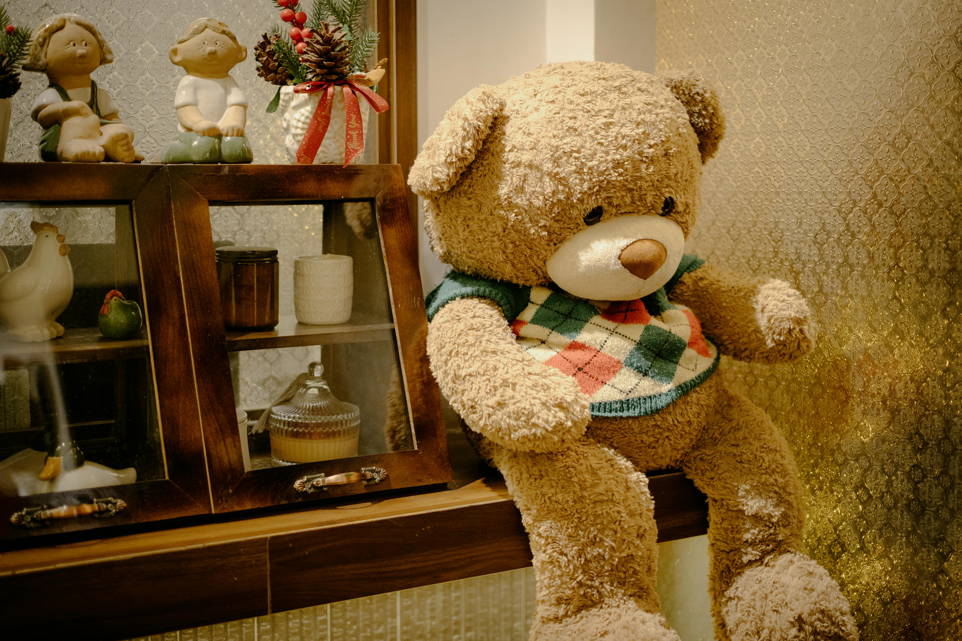 A fuzzy brown teddy bear wearing a checkered sweater sits beside a small wooden shelf with decorative figurines.