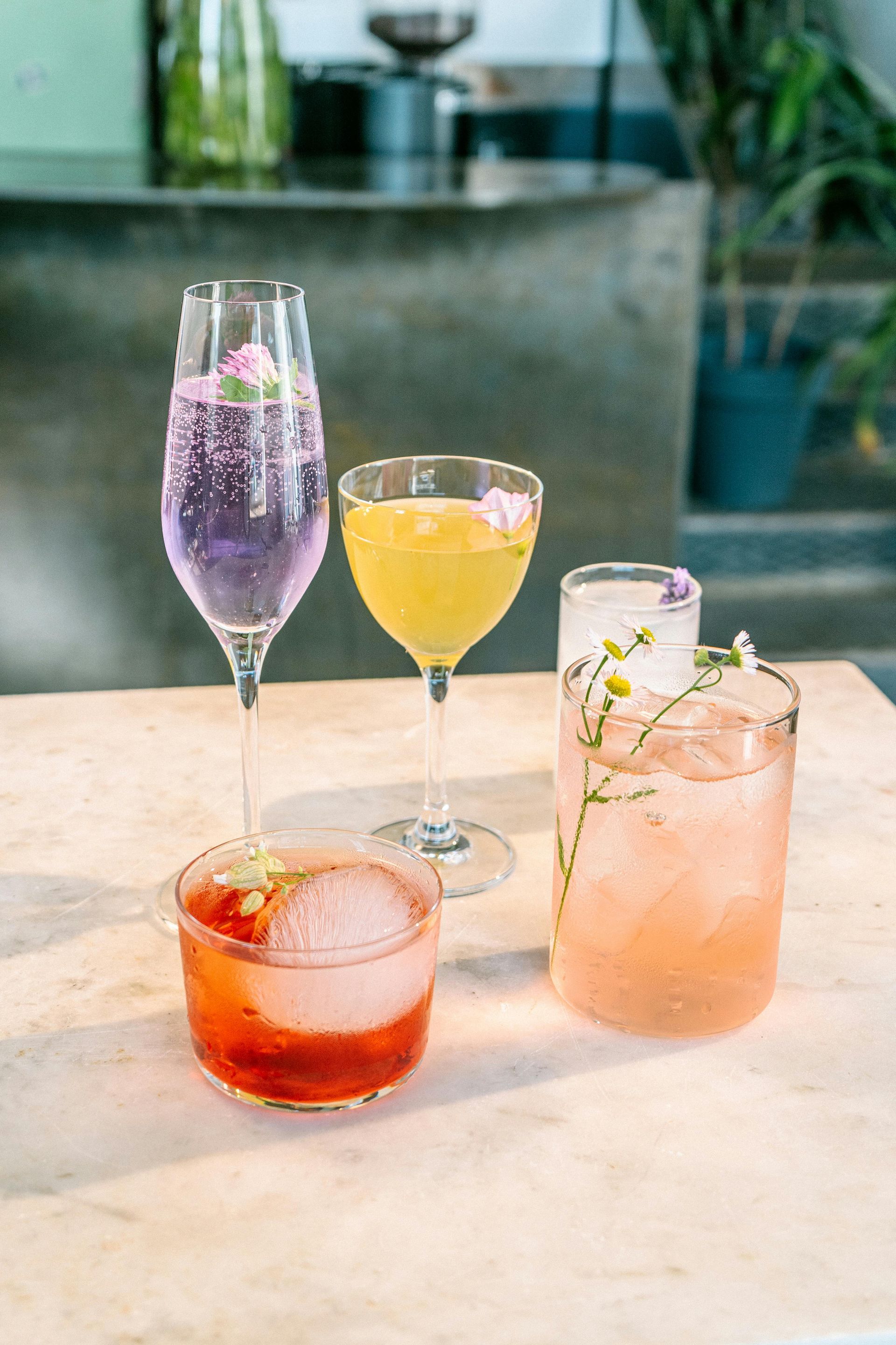 Four colorful cocktails in various glasses sit on a marble table in a softly lit, modern bar setting.