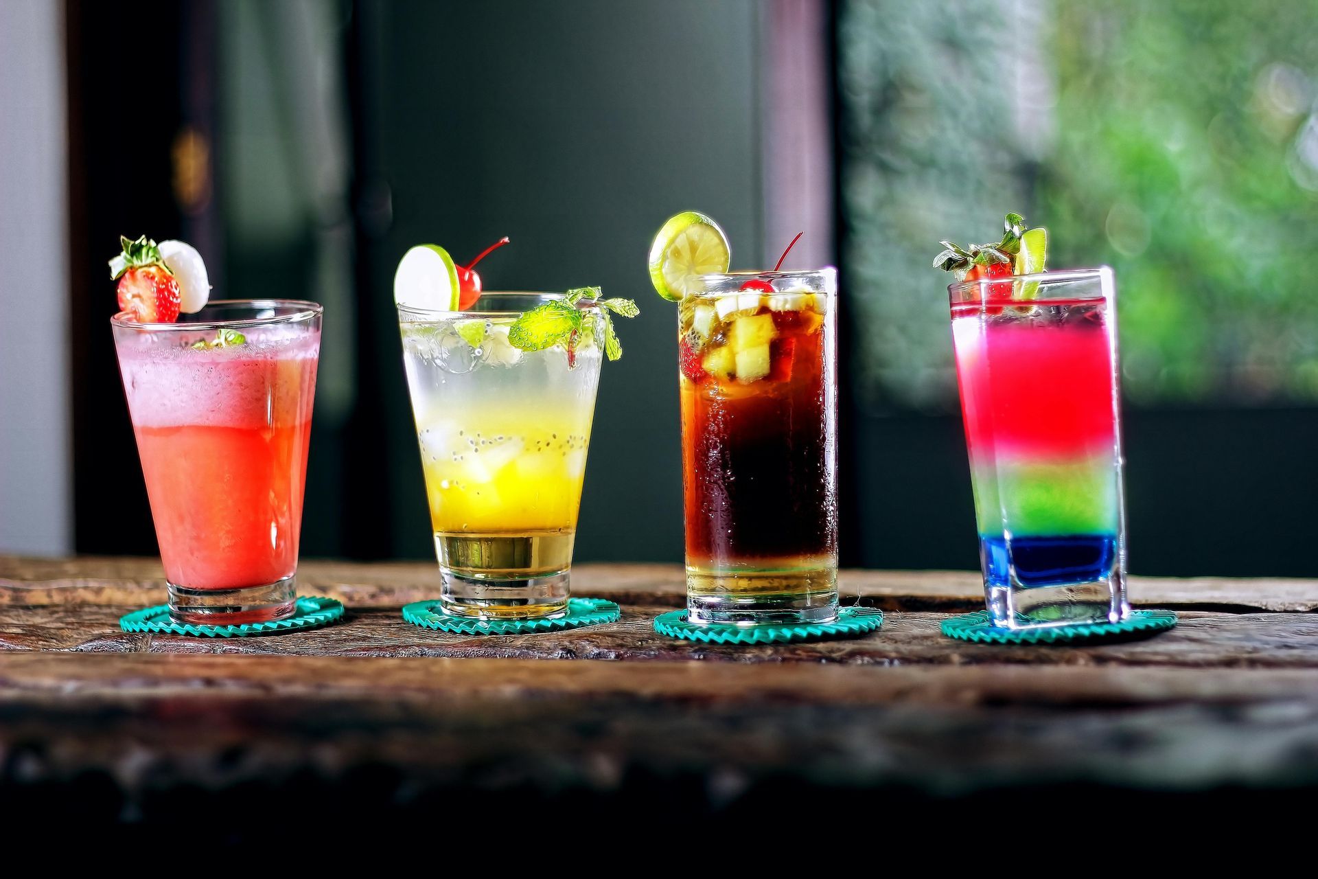 Four vibrant cocktails lined up on a rustic wooden table, each decorated with fresh fruit and resting on a small coaster.