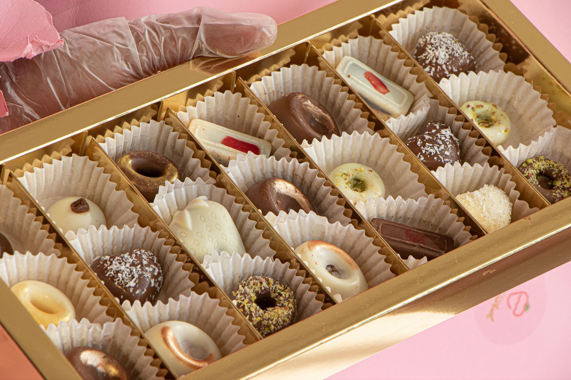 A box filled with assorted chocolates in paper liners, with a gloved hand holding the lid open against a pink background.