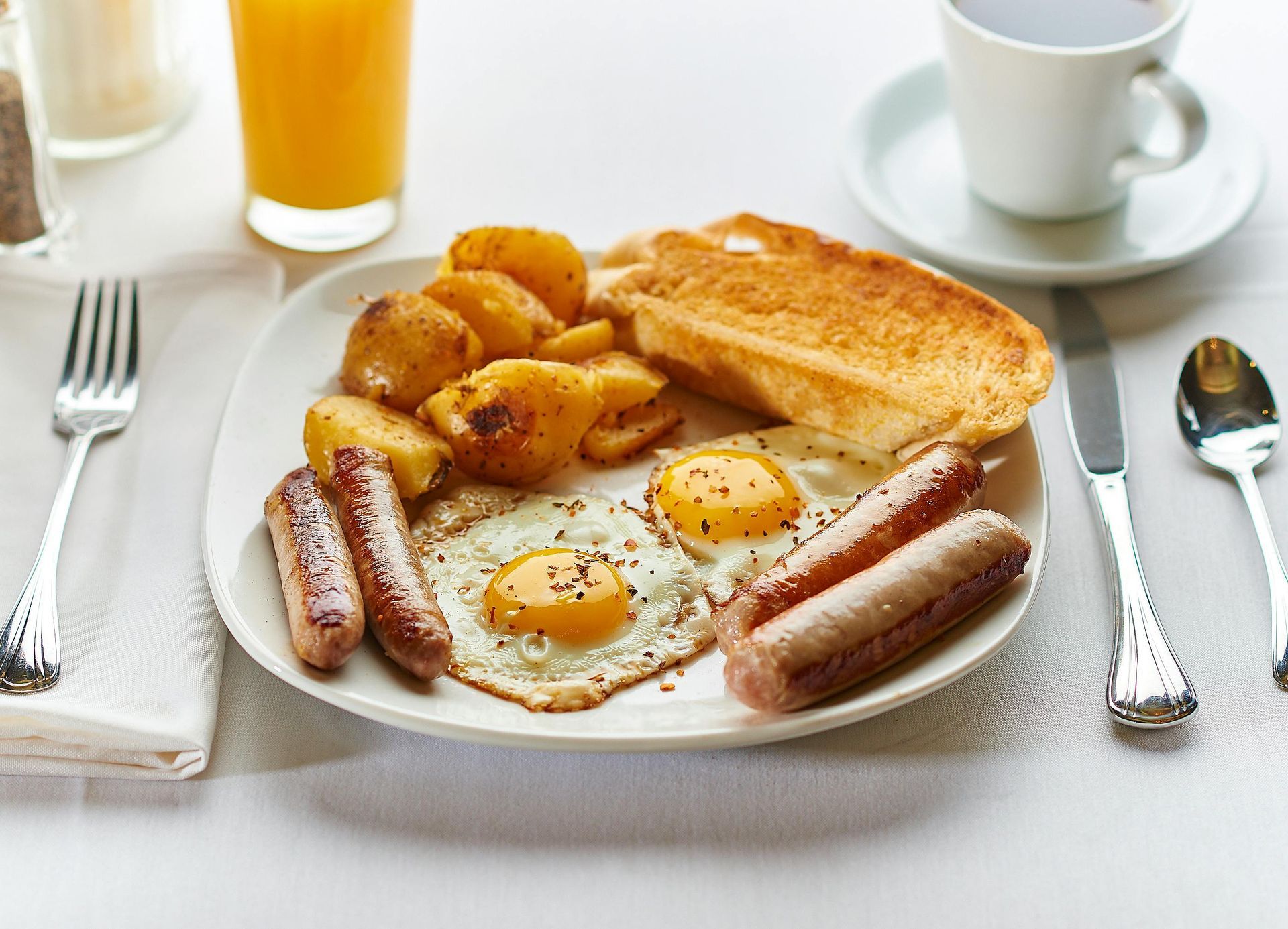A plated breakfast featuring two fried eggs, sausages, roasted potatoes, and toast, with juice and coffee on the side.