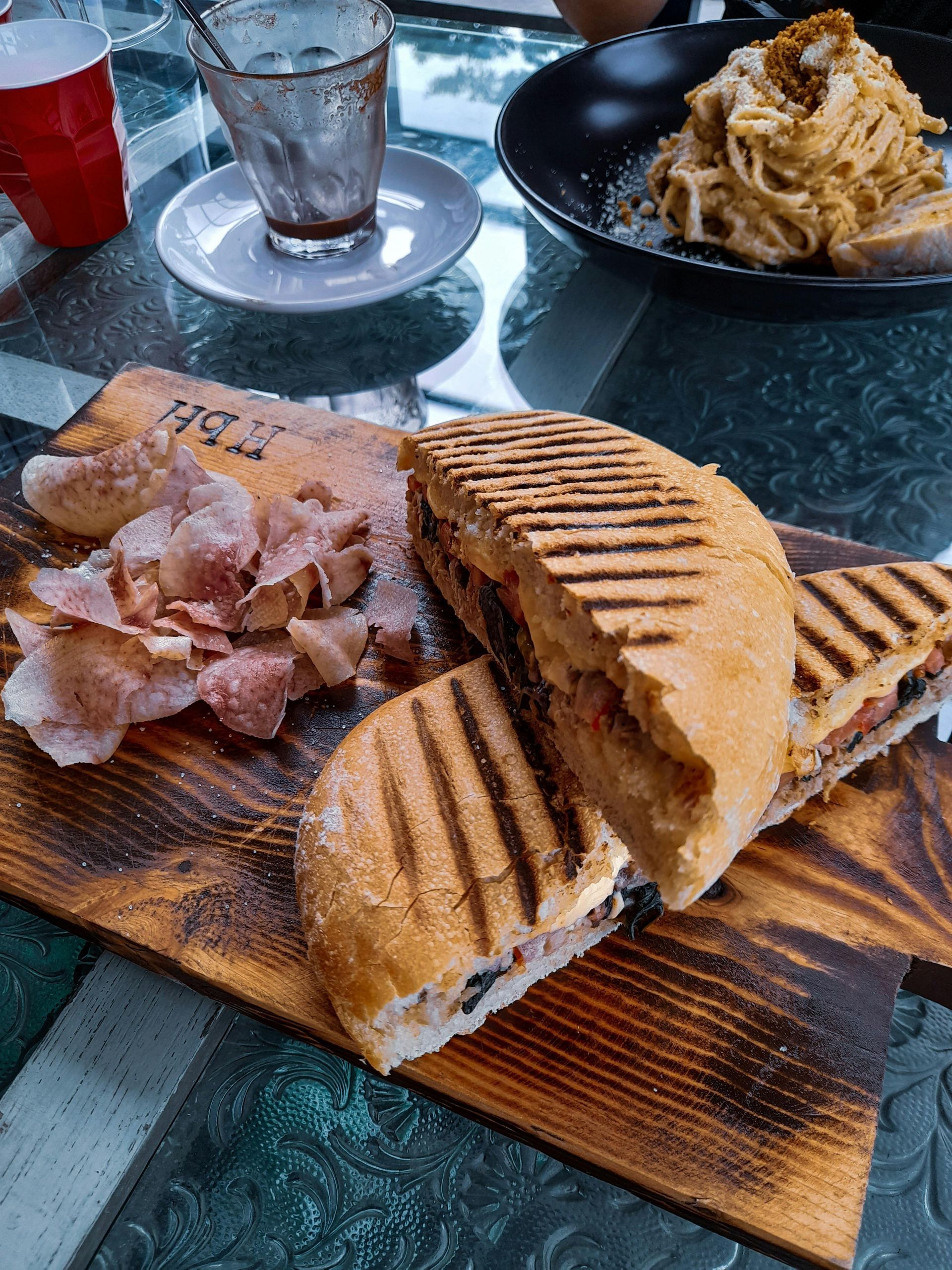 A wooden serving board features a grilled panini sandwich and a side of potato chips, with pasta and drinks nearby.