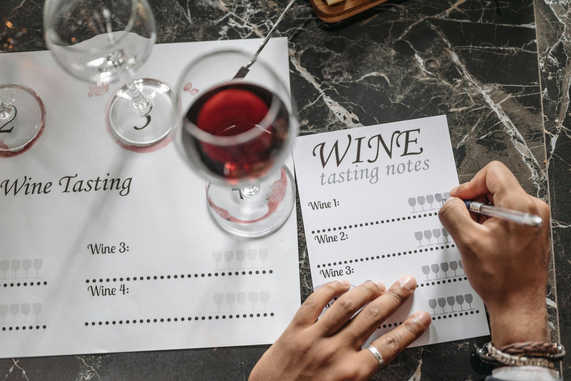 A person fills out a wine tasting note card with glasses of red wine on a marble table.