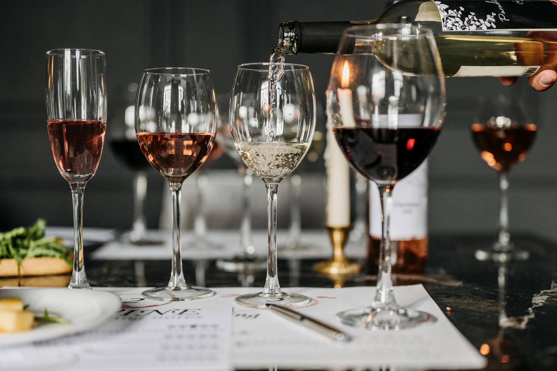 A hand pours white wine into a glass at a tasting event, with several glasses of red, white, and rosé wine on a table.