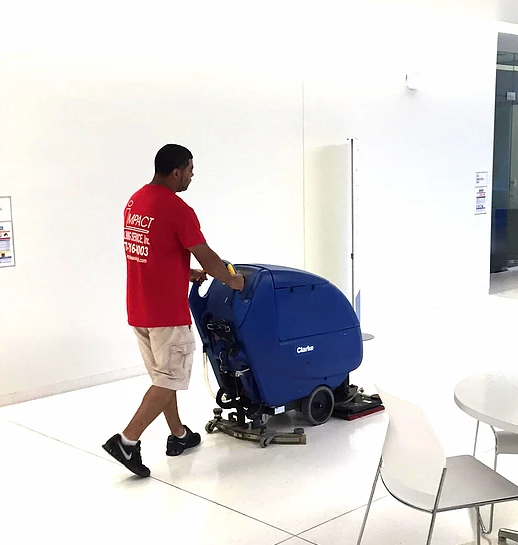 Man in red shirt operating a blue floor cleaning machine indoors. White walls, floor, and furniture in view.