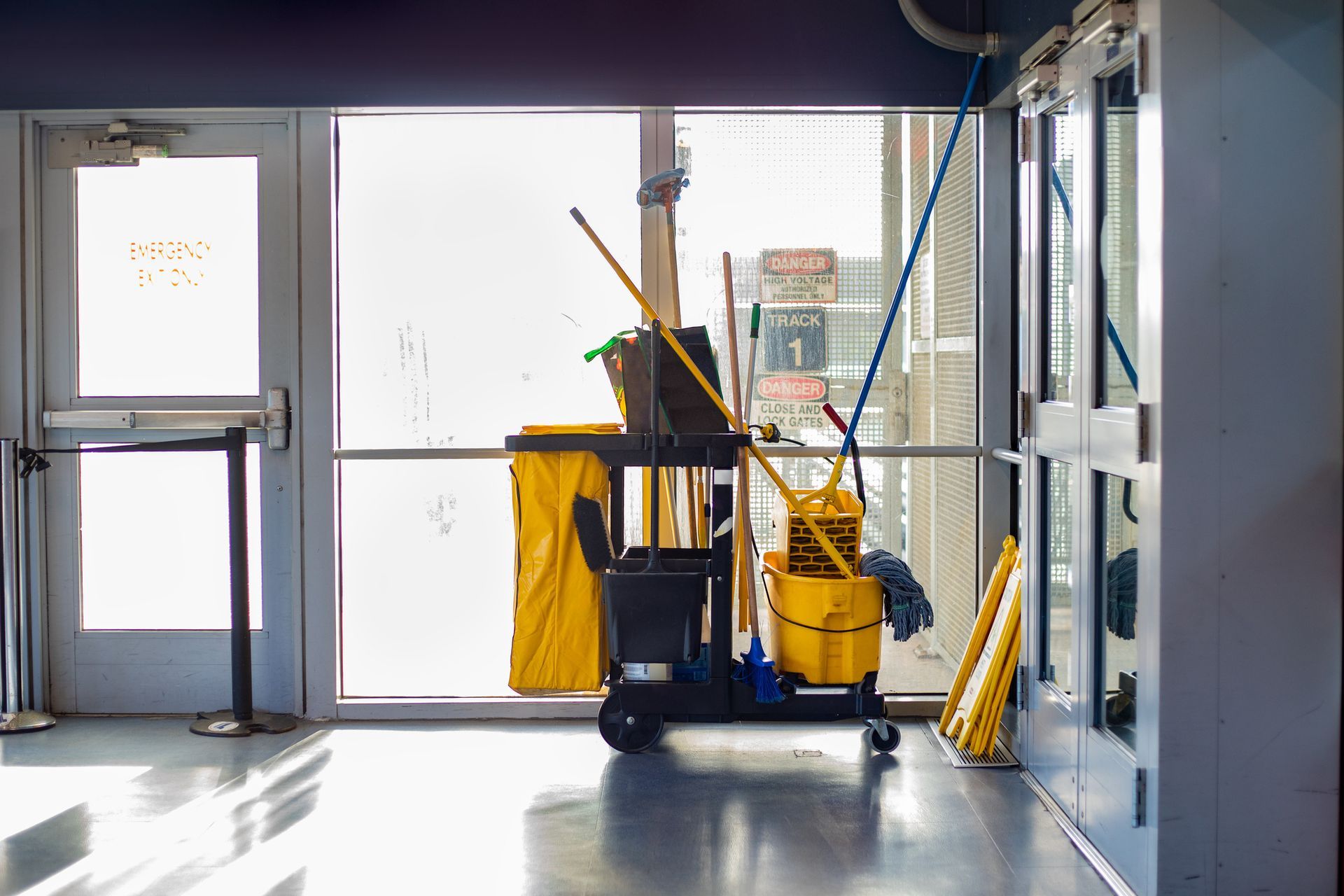 A cleaning cart with various supplies stands near a large window in a building