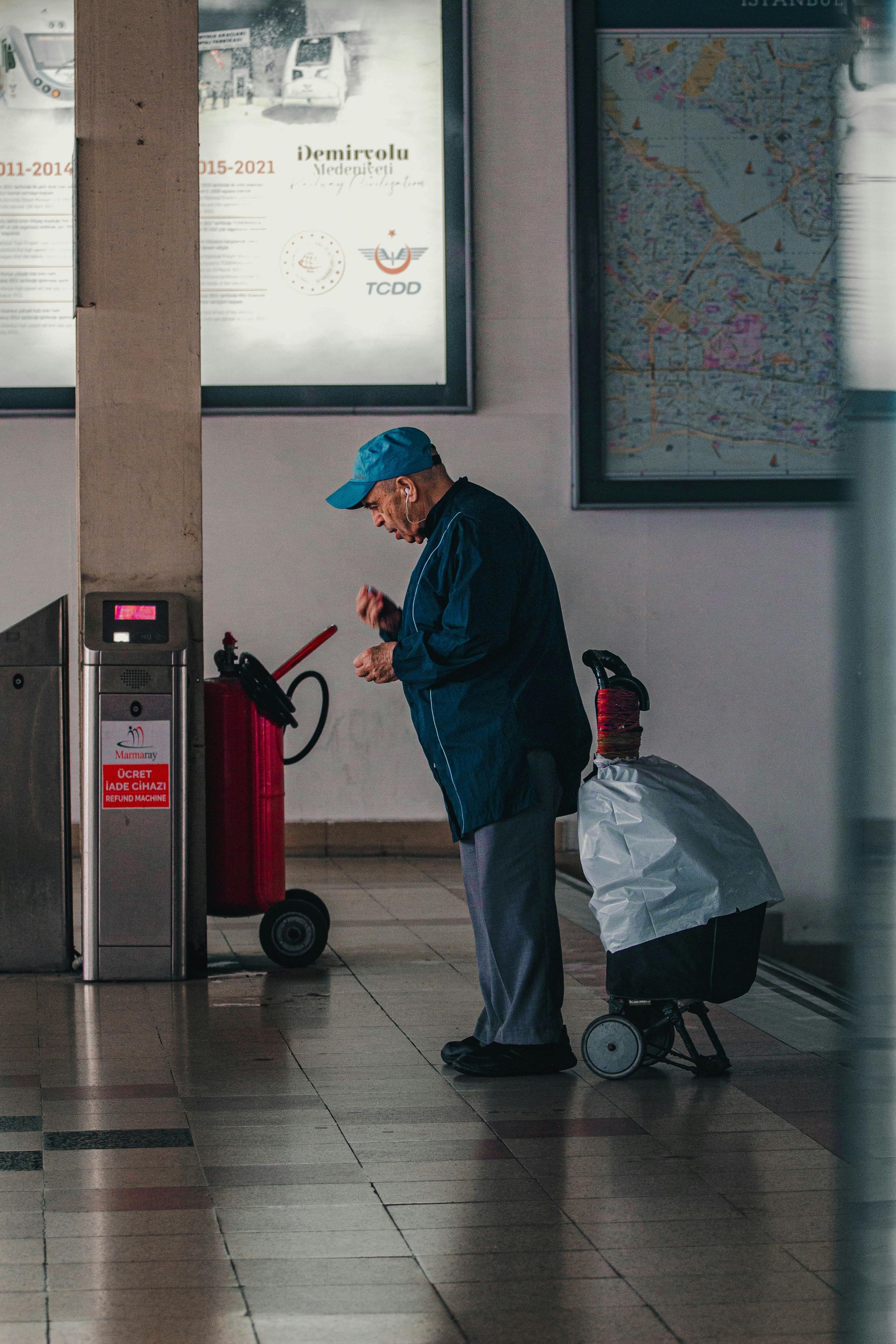 Elderly person with a blue hat and shopping cart at a train station, looking at something in hand.