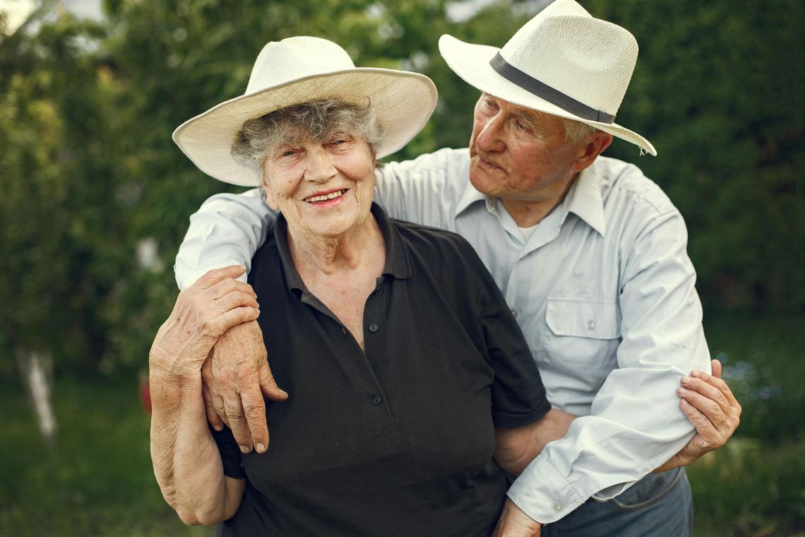Elderly couple in hats, the man embracing the smiling woman outdoors.