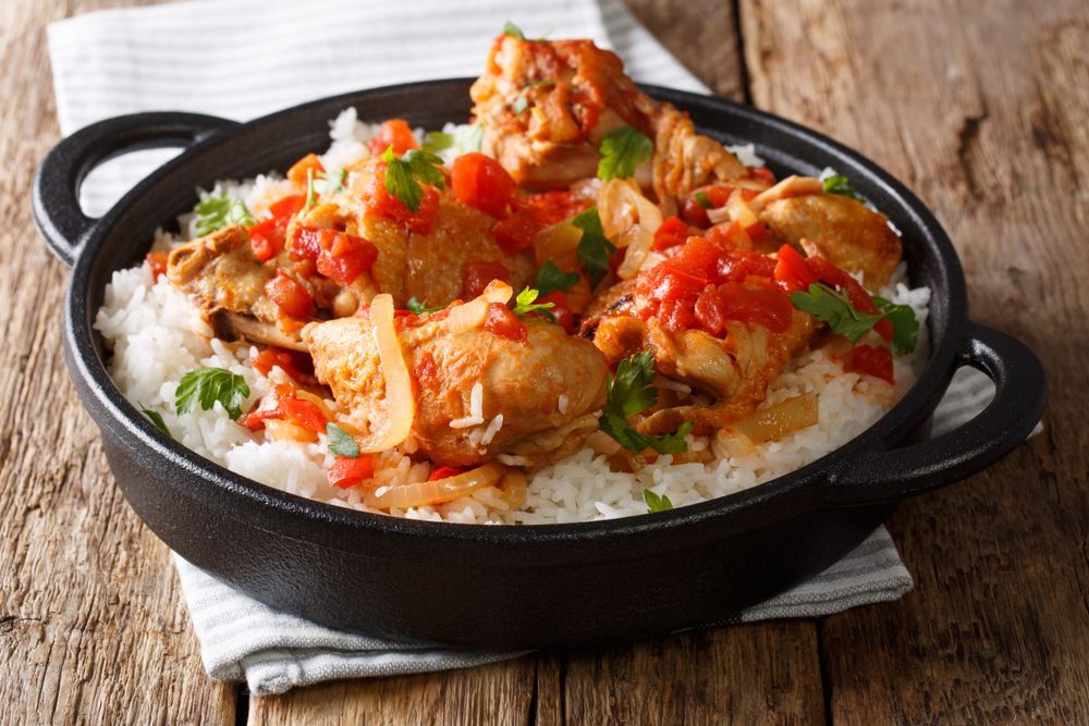 Chicken stew with tomatoes and onions over rice in a black bowl on a wood surface.