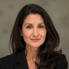 A professional headshot of a person with long dark hair, wearing a black blazer against a grey background.