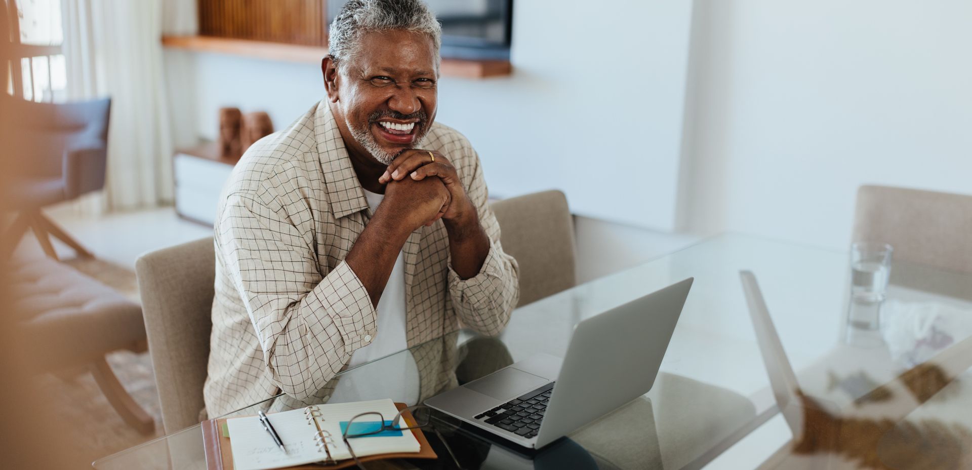 Smiling person with gray hair working on a laptop at a bright, modern dining table with an open notebook.