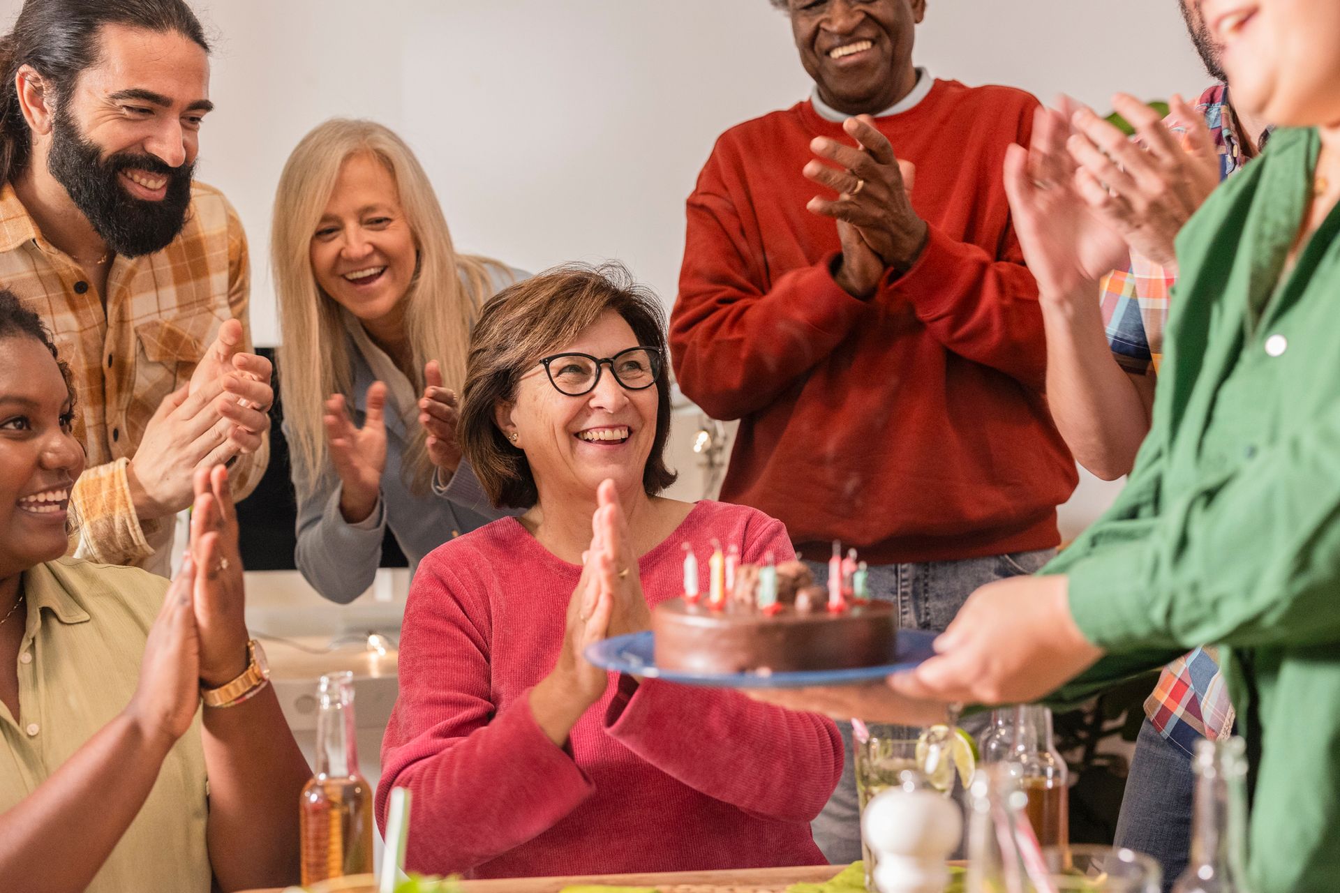 A group of people clapping as someone brings a chocolate cake with lit candles to a woman at a festive gathering.