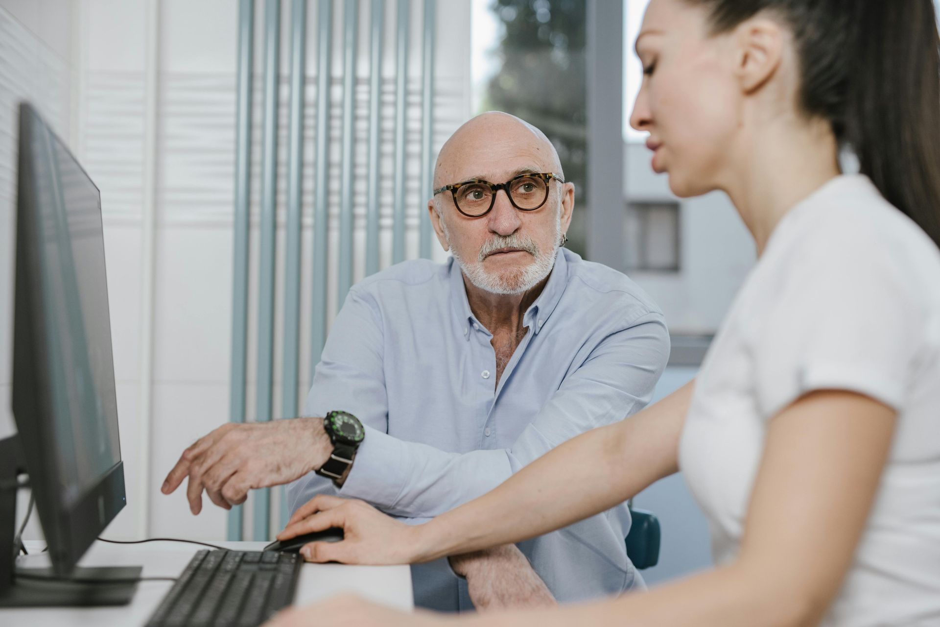 An individual points to a computer screen while looking at a colleague seated at a desk, collaborating in an office.