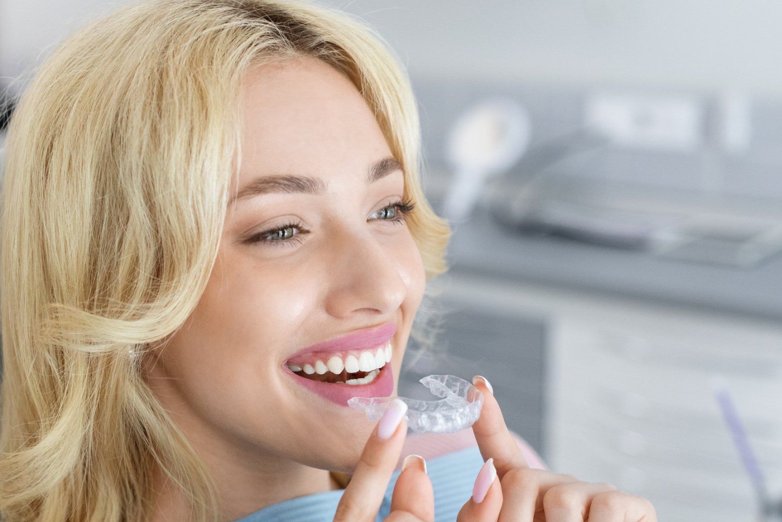 A woman is smiling while holding a clear retainer in front of her teeth.
