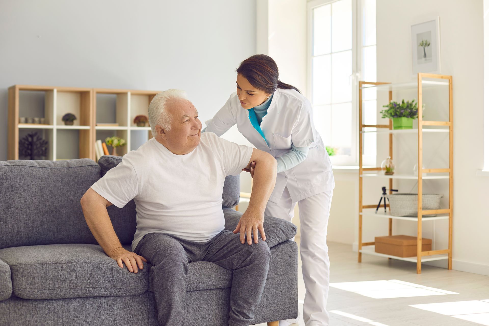 A nurse is helping an elderly man sit on a couch.