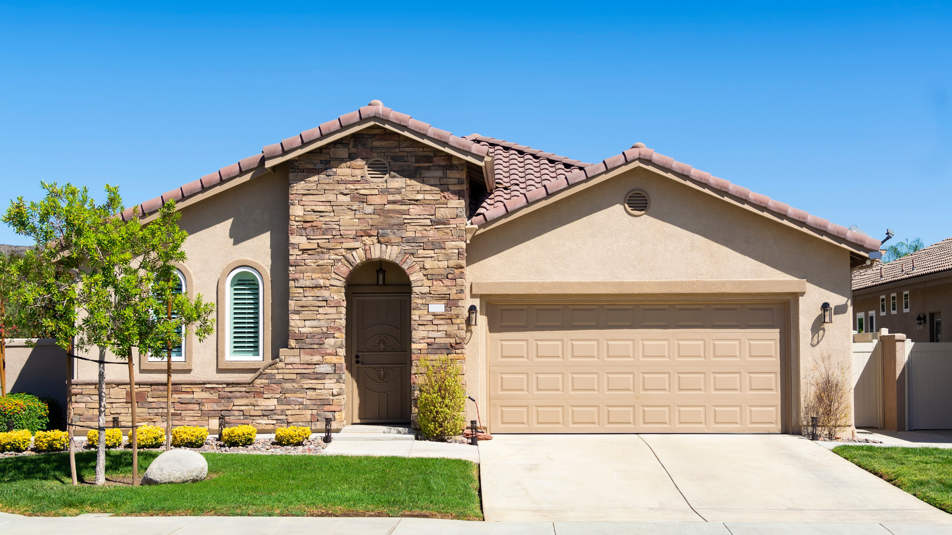 Tan stucco house with stone accents, garage, and lawn on a sunny day.
