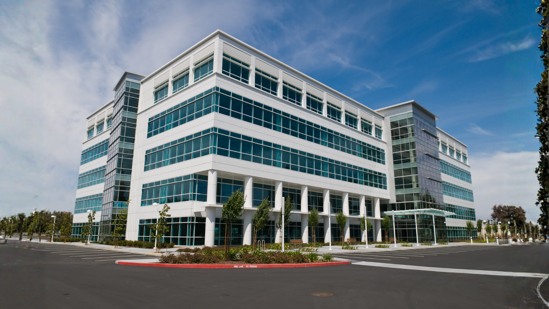 Modern white office building with blue-tinted windows under a blue sky, on a paved lot.