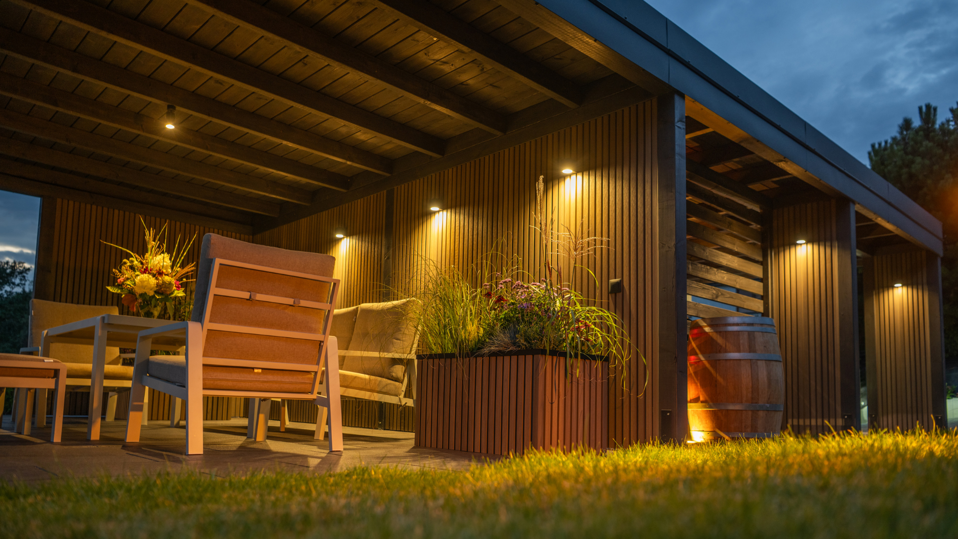 A modern outdoor pavilion at dusk with warm lighting, seating, and a large wooden barrel on a grassy lawn.