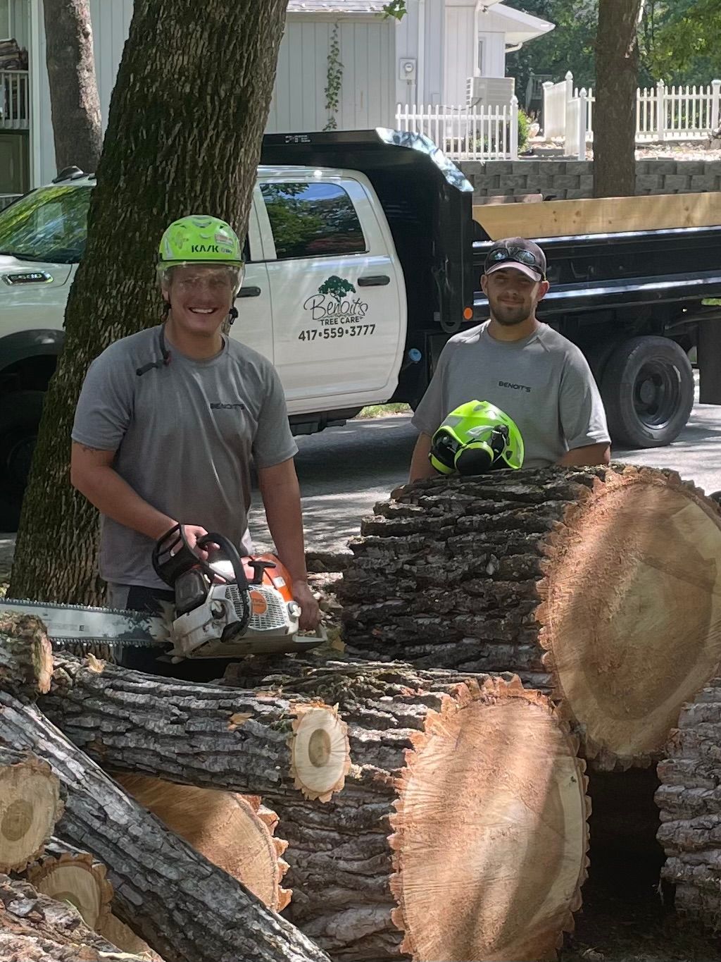 Two tree service workers with a chainsaw stand by cut logs. Truck in background.