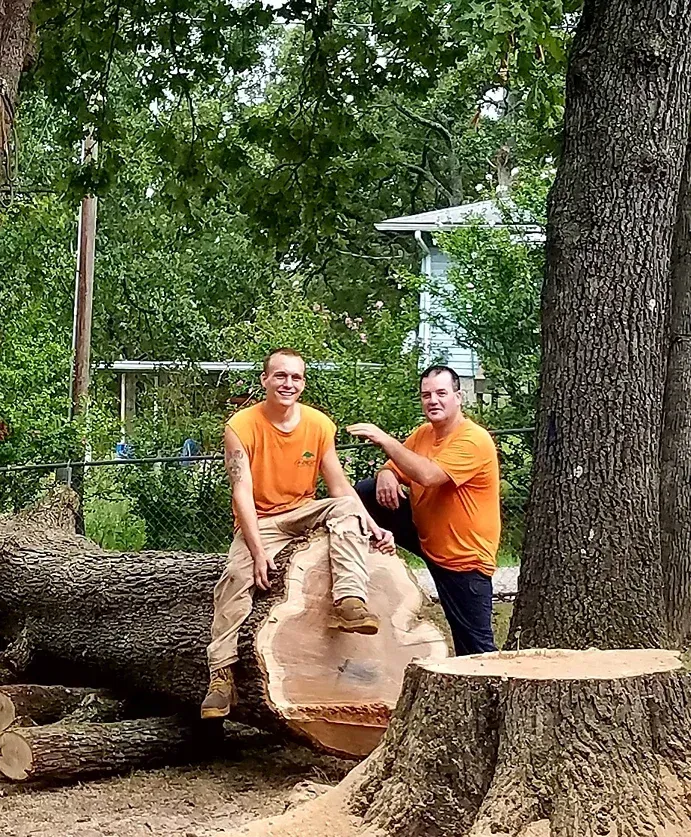 Two men in orange shirts pose near a large cut tree trunk outdoors; one sits on the log, the other points.