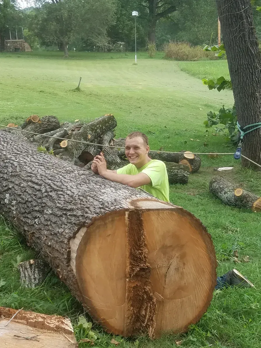 Man leaning on large cut tree trunk, smiling outdoors on green grass.
