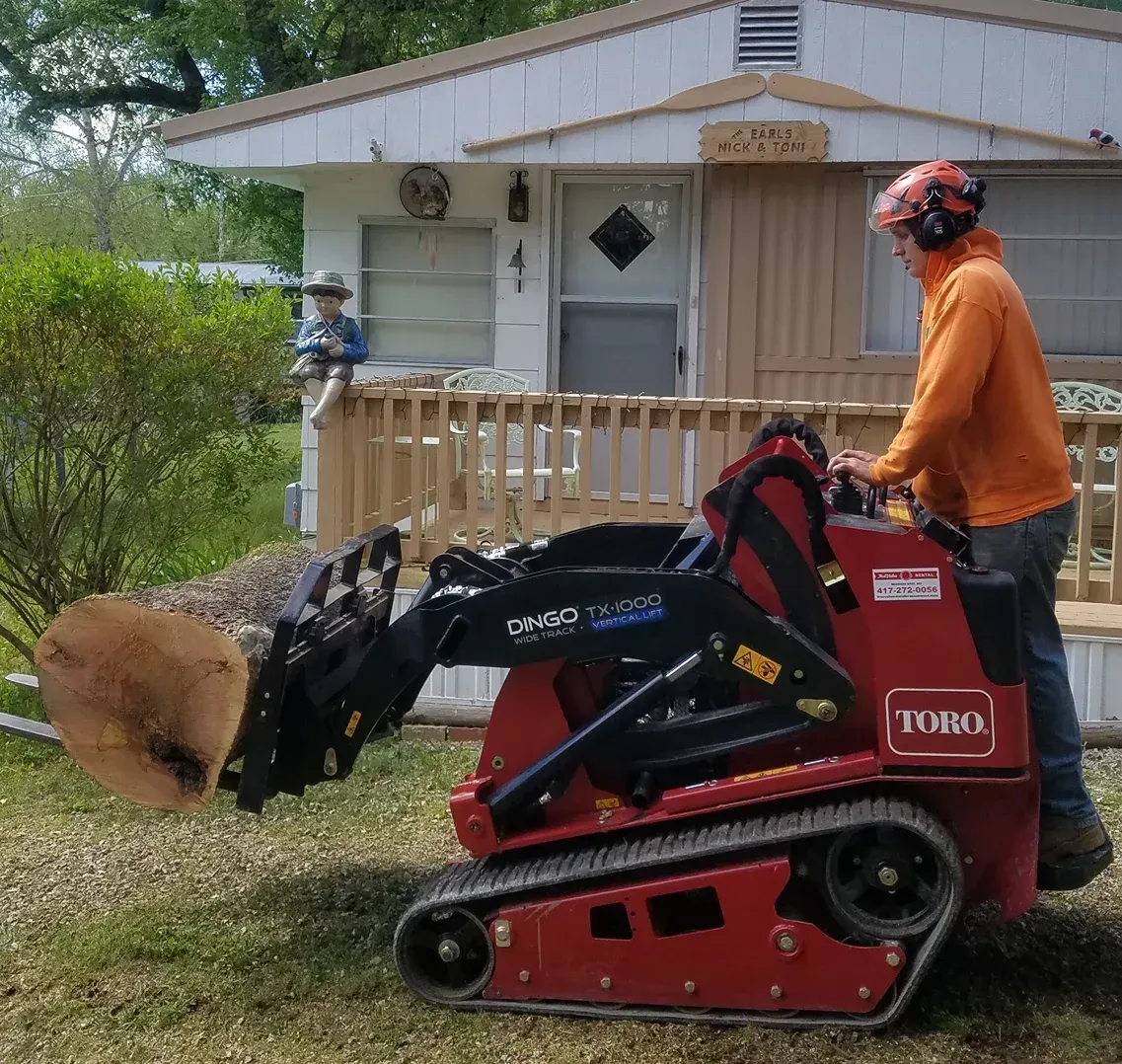 A man in orange operates a red Toro mini skid steer carrying a tree section near a house with a porch.