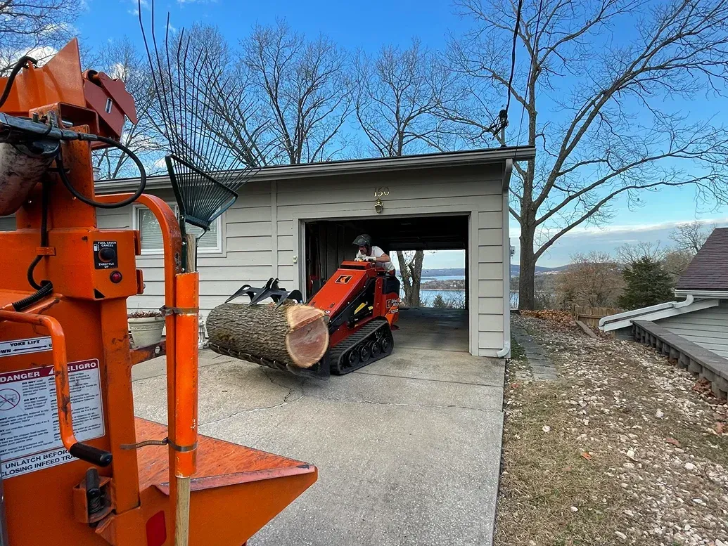 Mini excavator carrying a log towards a garage, with a wood chipper visible on the left.