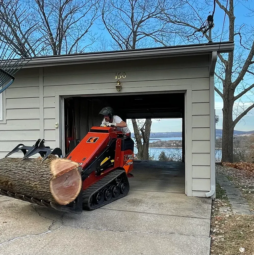 Man operates a mini-loader, carrying a large log into a garage; water and trees are visible in the distance.